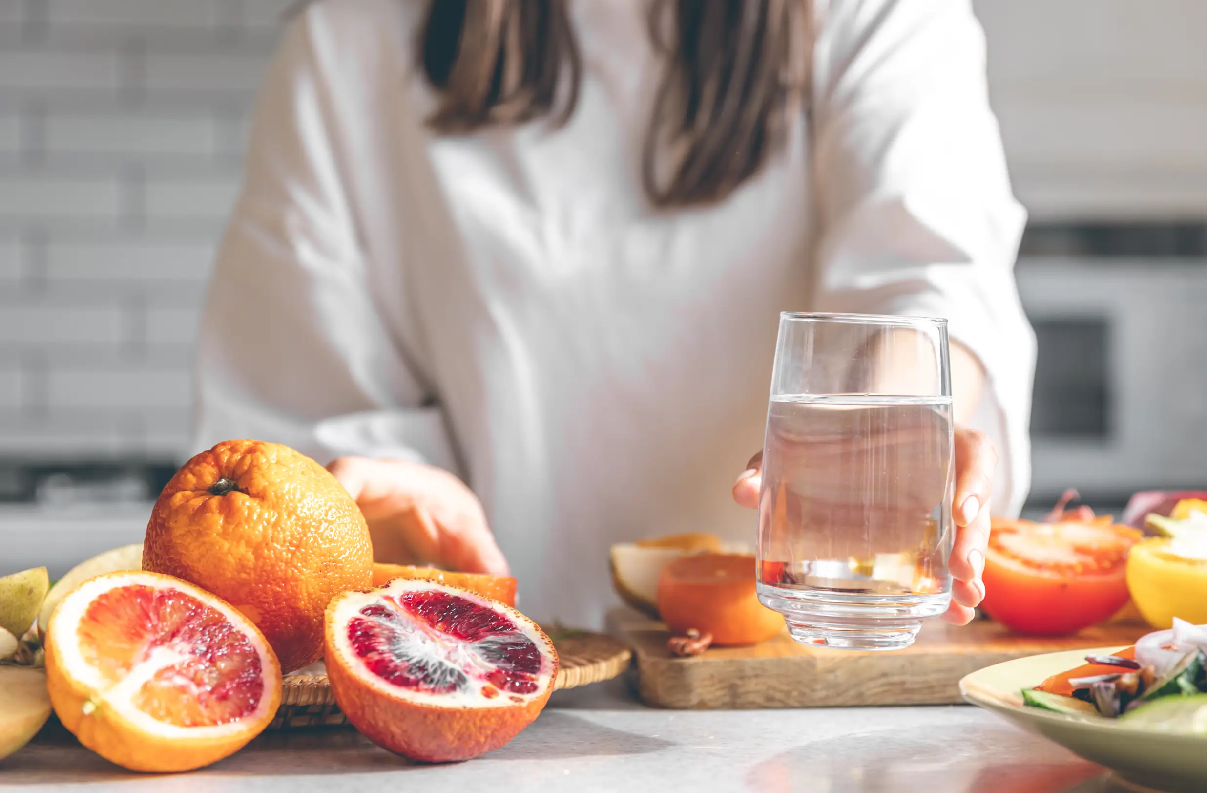 Woman holding a glass of water beside fresh citrus fruits and vegetables on a kitchen counter - IV Infusion