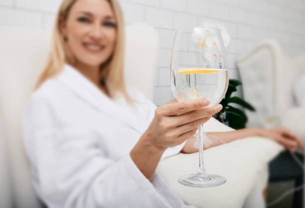 A smiling woman in a white bathrobe sits in a chair, holding a glass with a clear beverage and mint leaf, while a gloved hand (presumably a nurse or technician) tends to an IV line on her arm