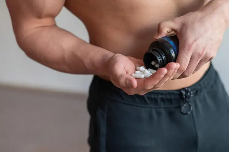 A muscular man is pouring white capsules from a black bottle into his hand.